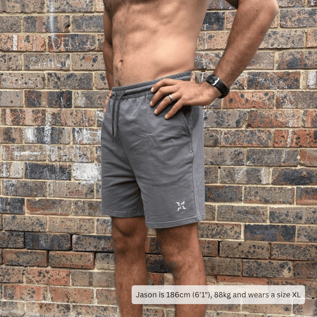 A man wearing gray athletic shorts stands against a weathered brick wall. The shorts are a medium gray color with a small white logo on the lower left leg.