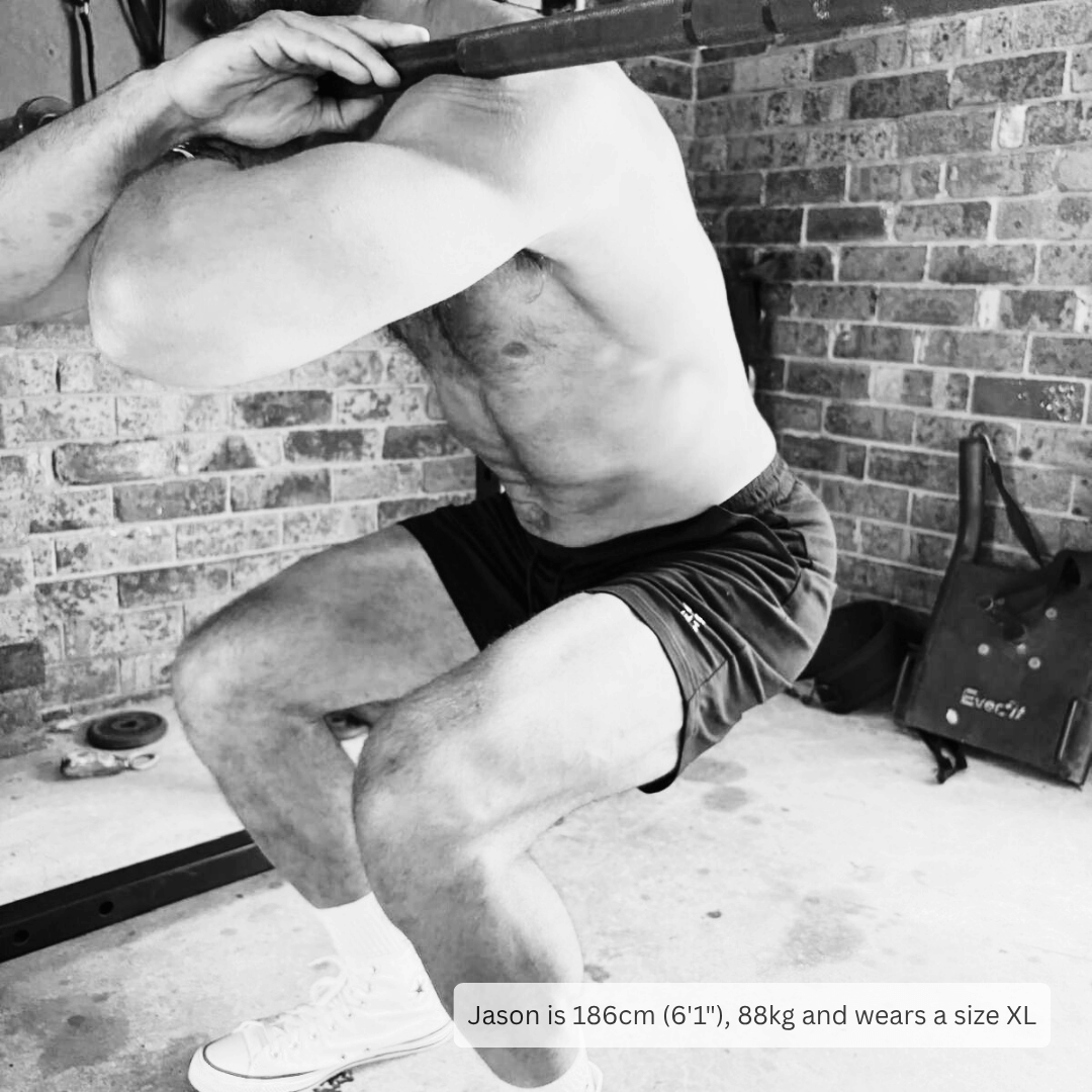 A black and white lifestyle photo of a man performing a squat in a garage gym setting, wearing dark athletic shorts and white sneakers. A text overlay at the bottom provides sizing information for the shorts.