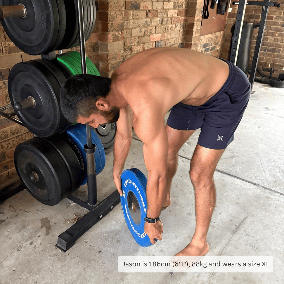 A shirtless man in navy athletic shorts bends down to place a blue weight plate on a rack in a gym with a brick wall background. Text at the bottom reads: 'Jason is 186cm (6'1''), 88kg and wears a size XL'.