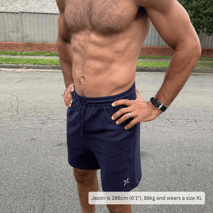 A man wearing navy blue athletic shorts, standing outdoors on a paved street. The shorts have a drawstring waistband and a small white logo near the lower left leg.